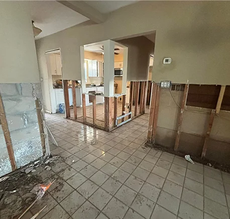 Water-damaged home interior with removed walls, exposed studs, debris on tiled floor, and a visible kitchen area.