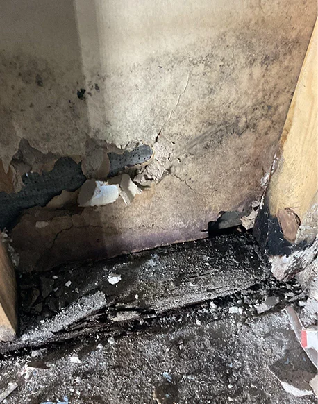 A close-up view of the corner of a room showing severe fire damage. There is black, charred debris on the floor, and the paint on the wall above is peeling and stained with soot.