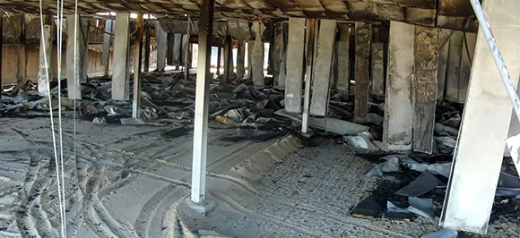 The severely burnt-out interior of a commercial building, showing charred roof trusses and support beams, with the floor completely covered in ash and debris from a fire.