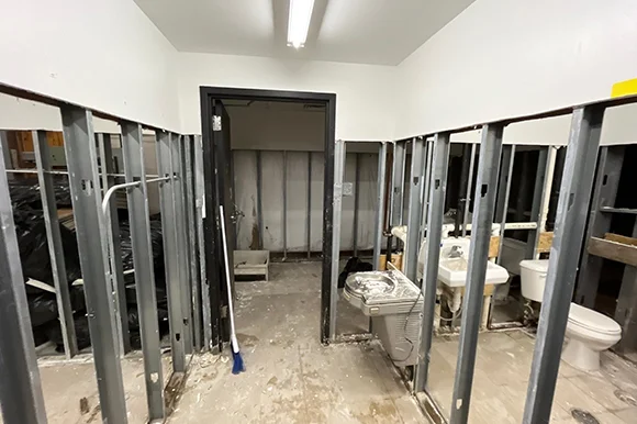 A commercial restroom or similar area under construction or restoration, with exposed metal studs, black plastic sheeting on the left, and multiple toilets and sinks removed or in disarray on the right.