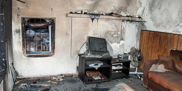 A fire-damaged room with blackened, soot-stained walls, a damaged television on a stand, and debris scattered on the floor.