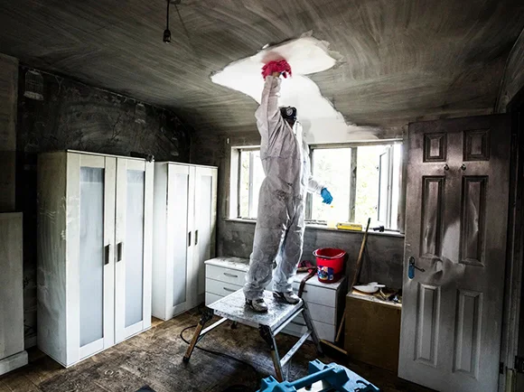 A restoration worker in a full white protective suit and red hard hat stands on a stepladder to clean the soot-damaged ceiling of a residential room.