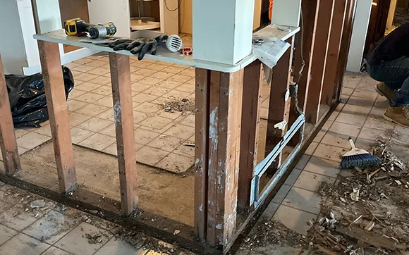 A room undergoing water damage mitigation, with wooden wall studs exposed and debris on the floor. There are tools like a drill and a broom visible.
