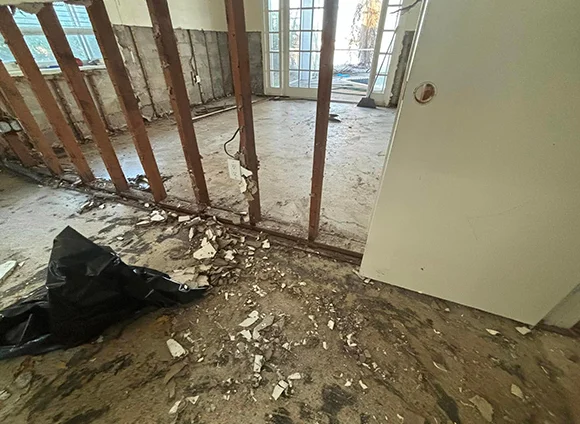 Water-damaged residential room with exposed studs, debris on floor, and French doors visible during remediation.